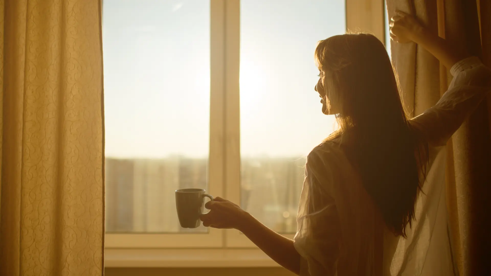 Woman standing by a sunlit window holding a cup of coffee, symbolizing the energizing physiological effects of coffee