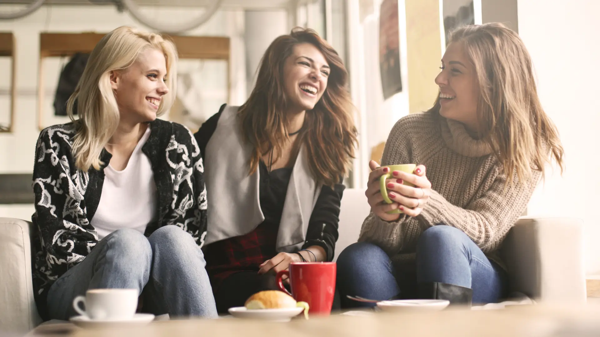 Three young women sitting on a couch, laughing and drinking coffee together, representing the social and psychological effects of coffee consumption